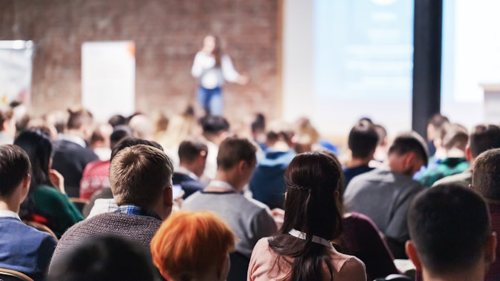 Adult people at conference listen to woman speaker providing lecture on scene in big conference hall.