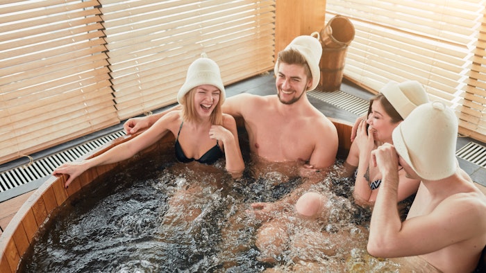 Group of male and female friends visiting bathhouse in holidays, being overjoyed and happy, enjoying jacuzzi in hot wooden round barrel