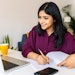 Young adult indian student woman taking notes while using laptop computer at home. Millennial ethnic female learning online listening virtual video call. Business and education concept.