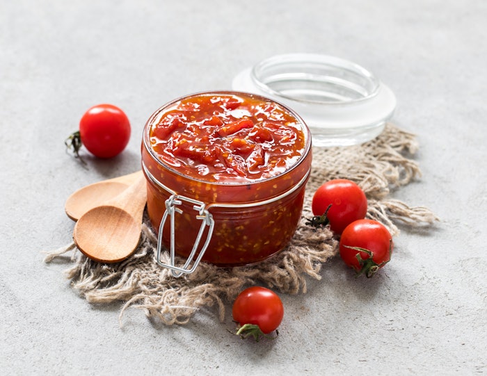 Cherry tomato jam in a glass jar on a linen napkin on a light gray background in rustic style