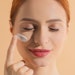 redheaded beautiful woman eyes closed applying soothing lotion to face beige background