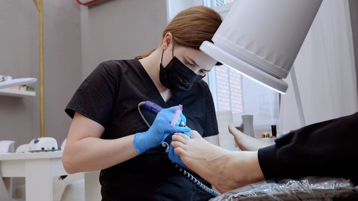 Podiatrist wearing black mask and blue gloves using electric nail drill for pedicure treatment on client's feet in beauty salon