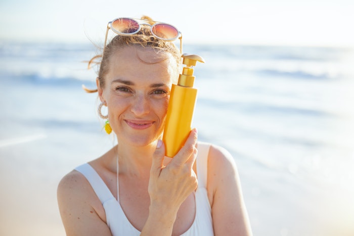Woman On Beach Holding Sunscreen Bottle Smiling Adobe Stock 561891115 (1)