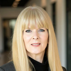Professional blonde woman in gray blazer and black top, smiling in office setting