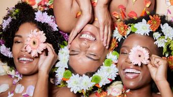 Three women smiling joyfully surrounded by colorful flowers including daisies, gerberas, and chrysanthemums in pink, white, and orange tones.