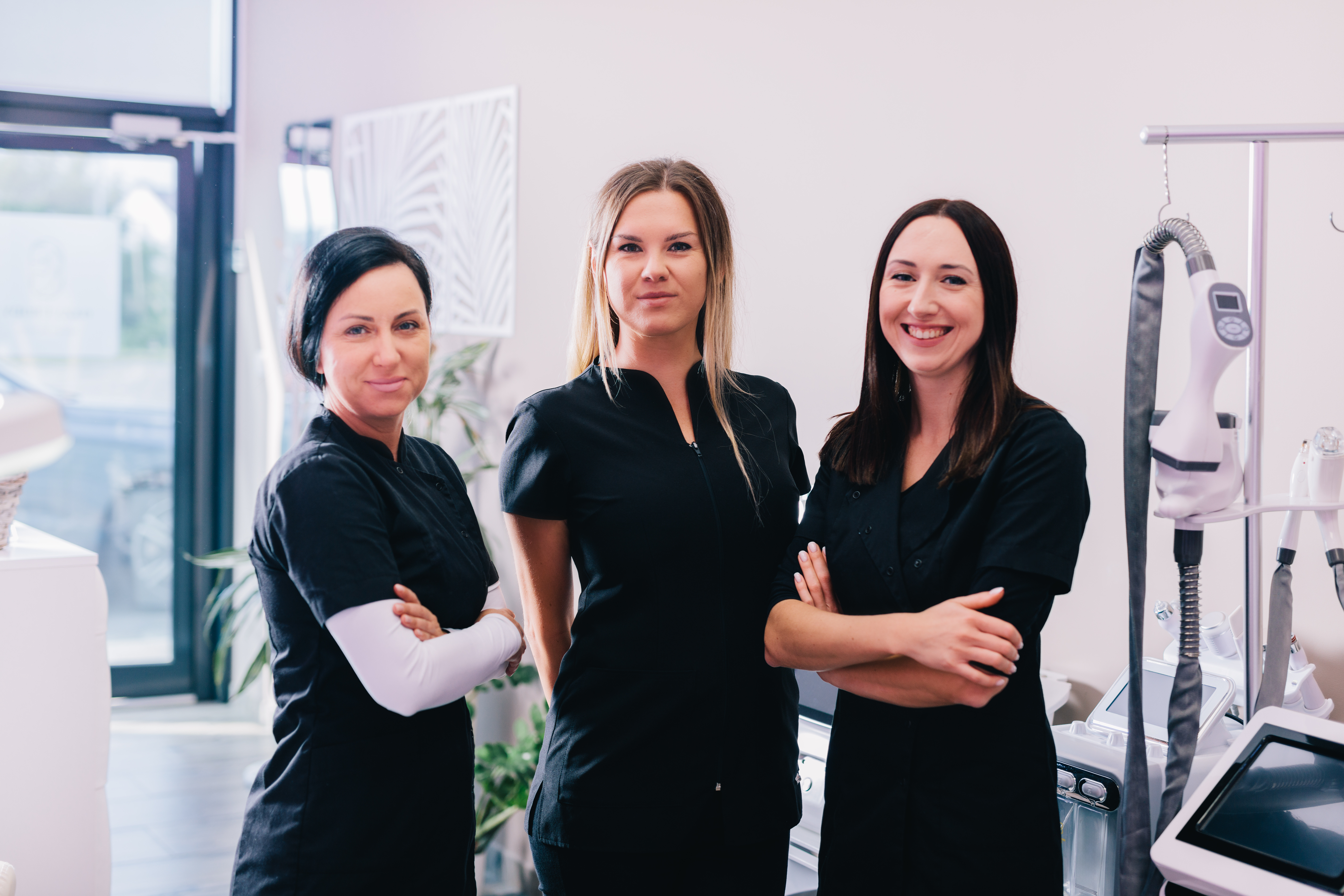 Three female healthcare professionals in black uniforms standing confidently in a modern medical clinic with aesthetic treatment equipment visible in the background.