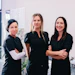 Three female healthcare professionals in black uniforms standing confidently in a modern medical clinic with aesthetic treatment equipment visible in the background.