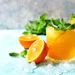 Three glasses of fresh orange juice with mint garnish and ice cubes, surrounded by orange slices on a light blue surface