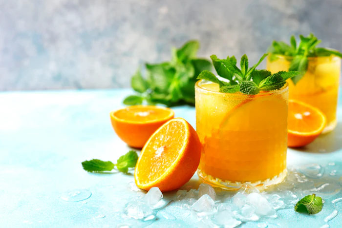 Three glasses of fresh orange juice with mint garnish and ice cubes, surrounded by orange slices on a light blue surface