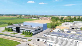 Aerial view of modern industrial facility with blue-roofed building, manufacturing structures, and surrounding agricultural fields