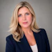 Professional headshot of a woman with blonde hair wearing a navy blazer and white top against a gray background