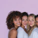 Four smiling young women posing together against a purple background in casual white clothing