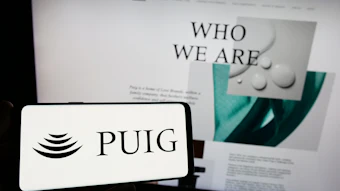 PUIG logo displayed on smartphone screen with 'Who We Are' website content visible in background