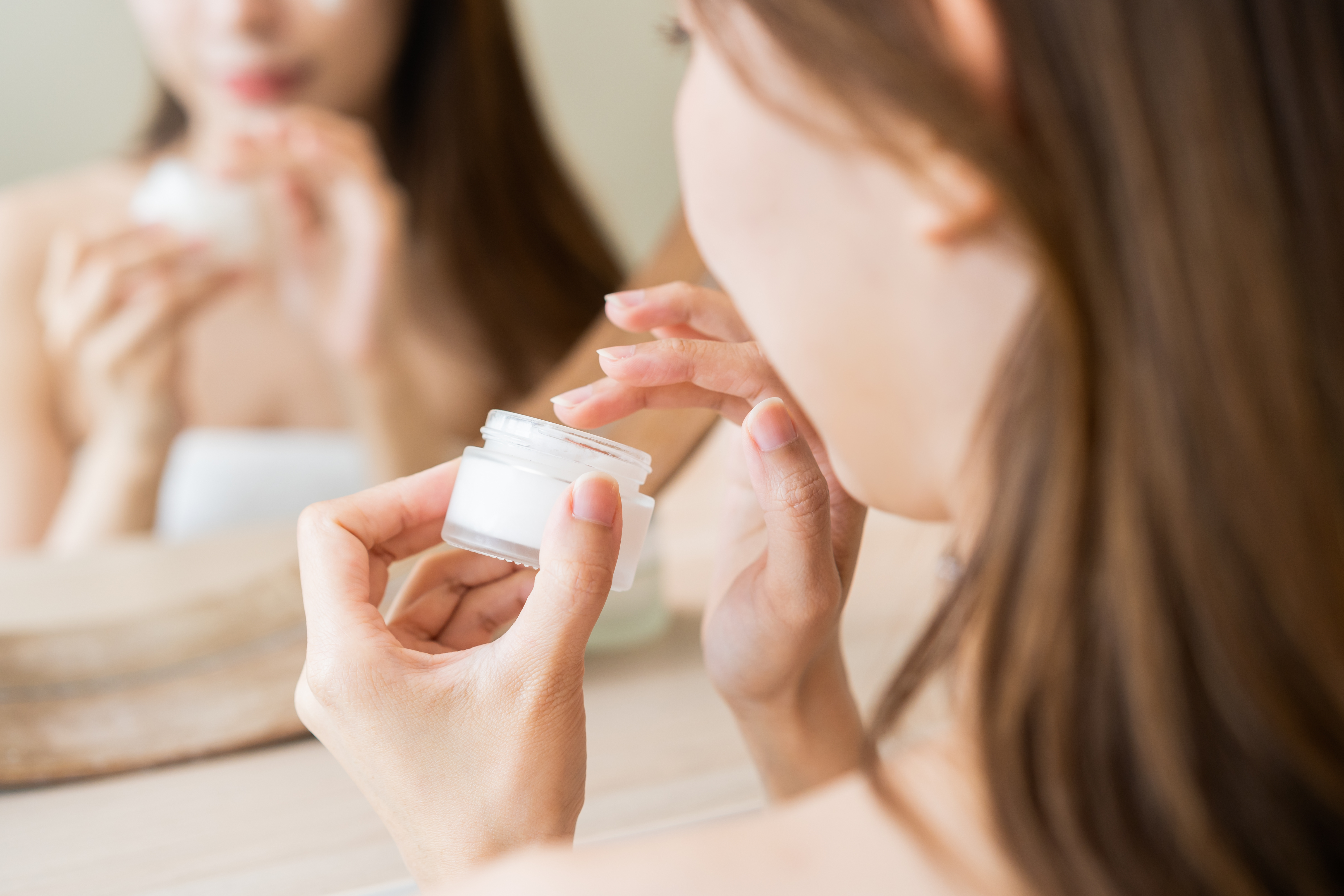 Woman applying facial cream from white jar with mirror reflection in background