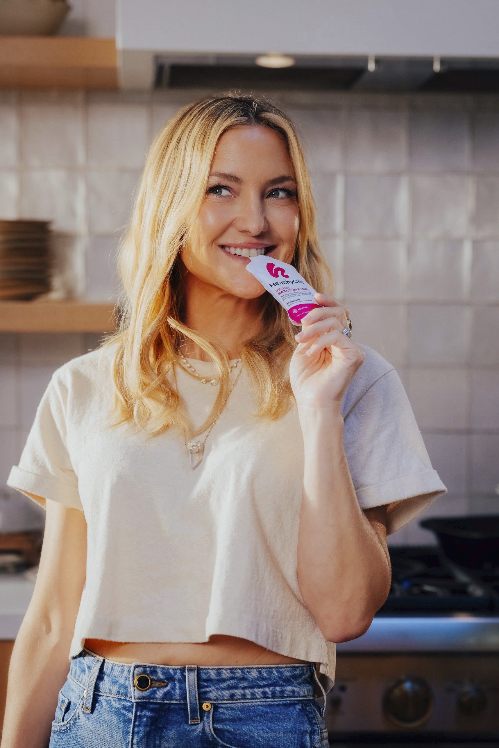 Smiling woman in white t-shirt holding pink cosmetic product in modern kitchen